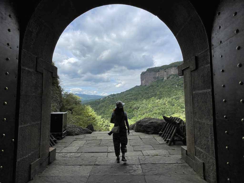 A human figure in the center of the images faces a stone archway. The archway looks out to cloudy skies and rolling hills.